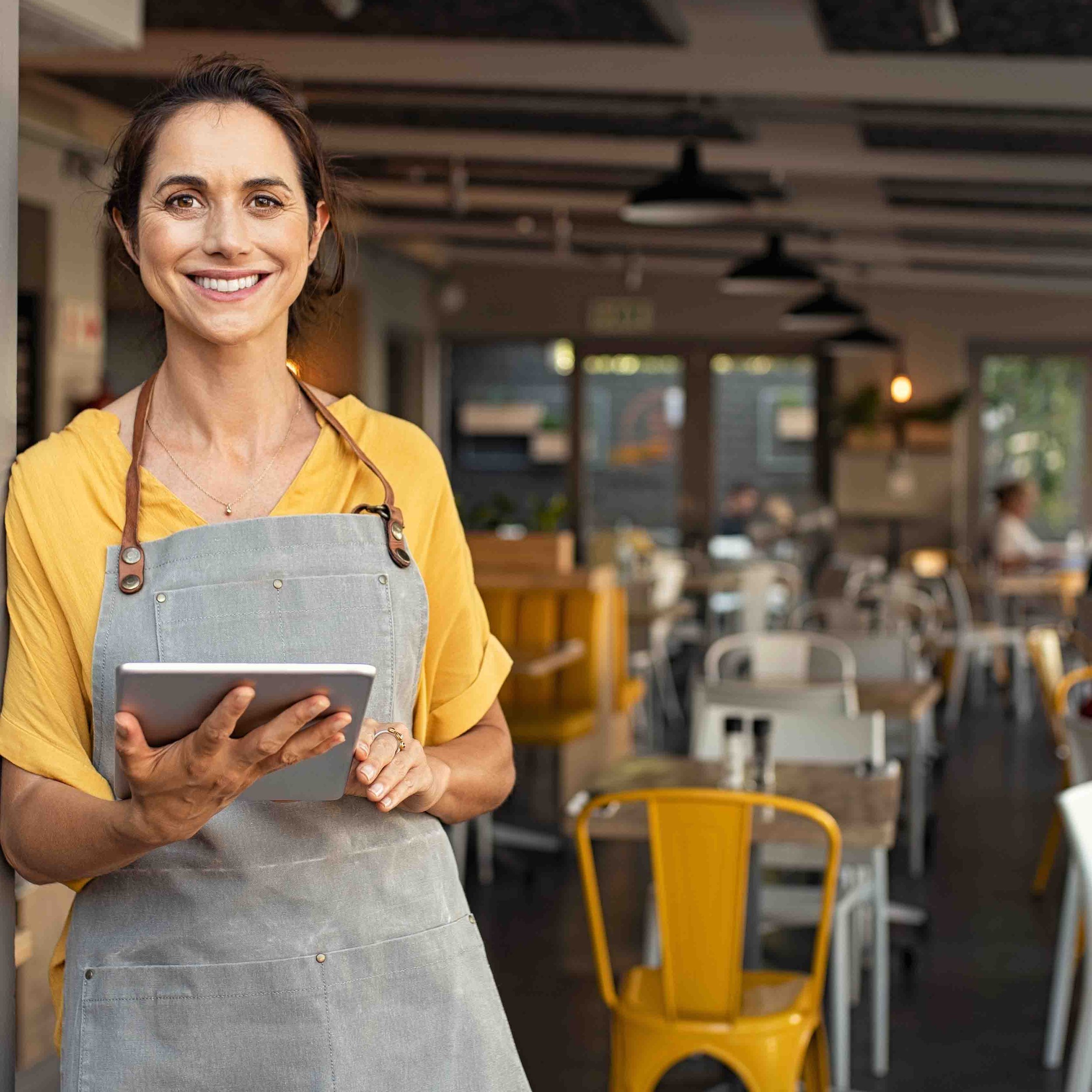 A woman stands in the doorway of her business with a tablet in her hand wearing an apron and a yellow top.