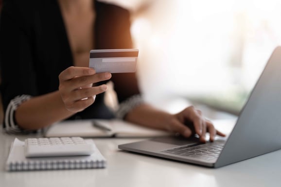 A person sits at a table with a computer, notepad, and calculator with a credit card in their hand.