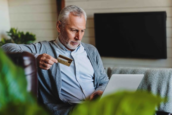 A man sits with a computer in his lap and a credit card in his hand.