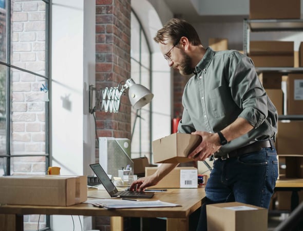 Man holding a box in a small warehouse checks his computer.