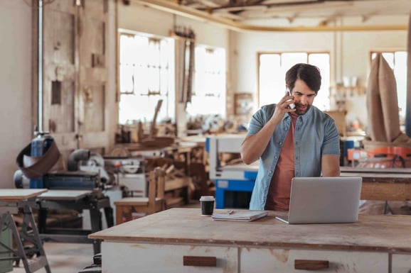 A man stands in a wood workshop at his work table on his computer open and talking on the phone.