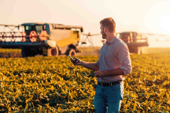 A man stands in a field with a large tractor behind him while he is on his phone.