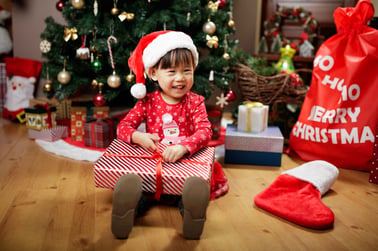 A child sits on the floor with a gift dressed in a Christmas outfit in front of the tree.