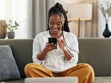 A female sits on the couch with laptop open in front of her while looking at her phone.