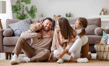 Parents and their 2 young kids sit on the floor in front of their couch playing together.