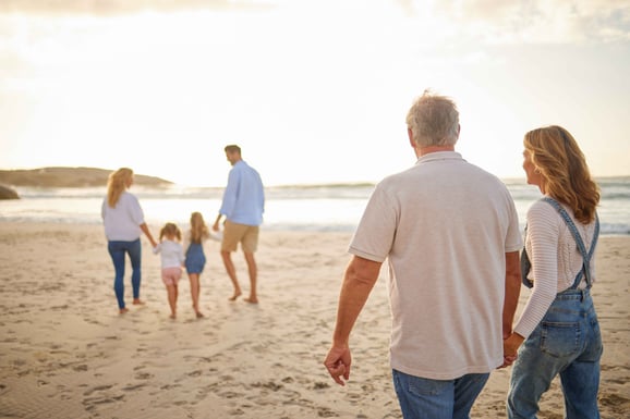 A couple walking on the beach looking at their grown children and grandchildren walking ahead.