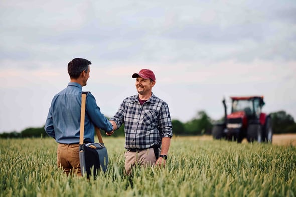 A farmer and a business man shake hands in a field with a tractor in background.