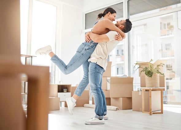 A man lifts a woman into the air in their home surrounded by boxes.