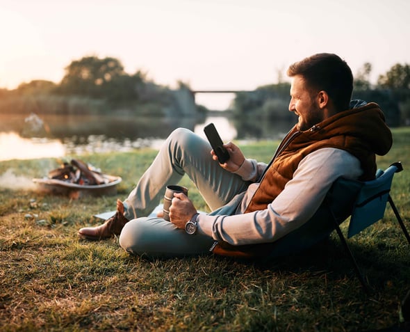 A man camping sits in a chair with a cup of coffee and is on his phone.