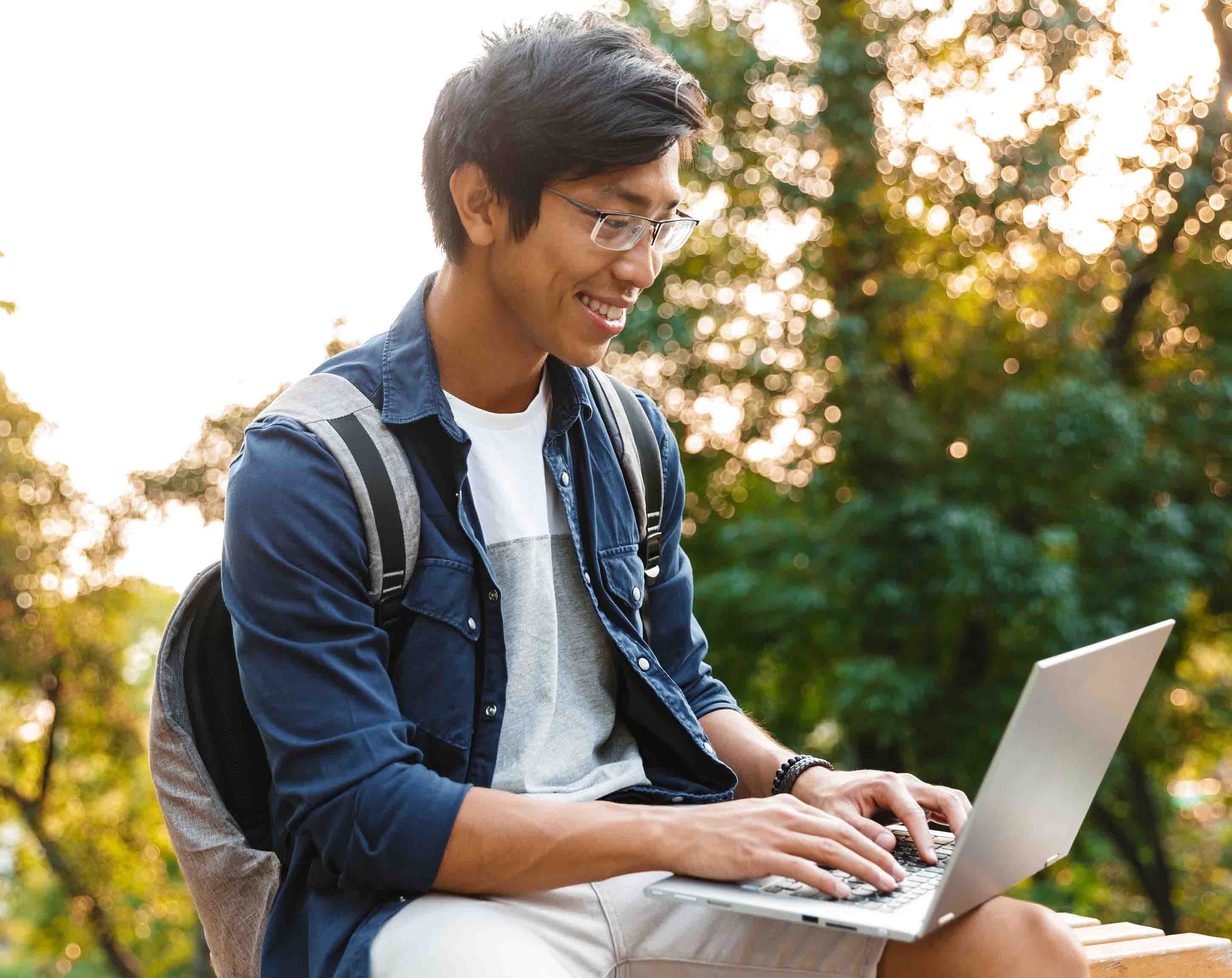 A younger man sits outside with a backpack on while working on his laptop.