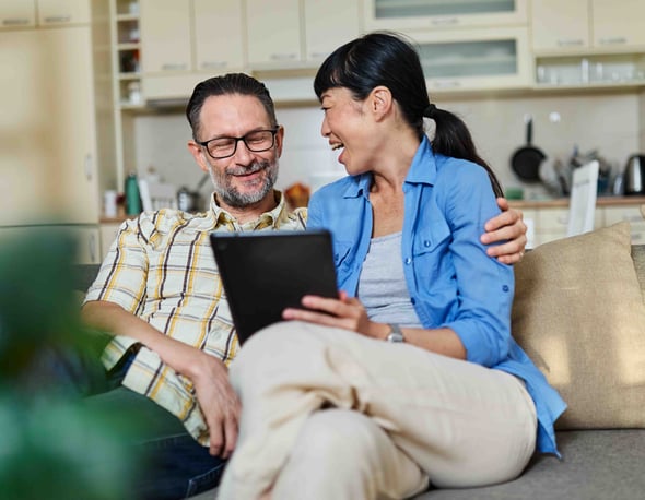 A man and woman sit on a couch together in their home with the woman holding a tablet.