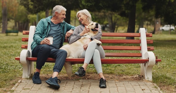 An older couple sit on a park bench together with a golden retriever sitting in between them.