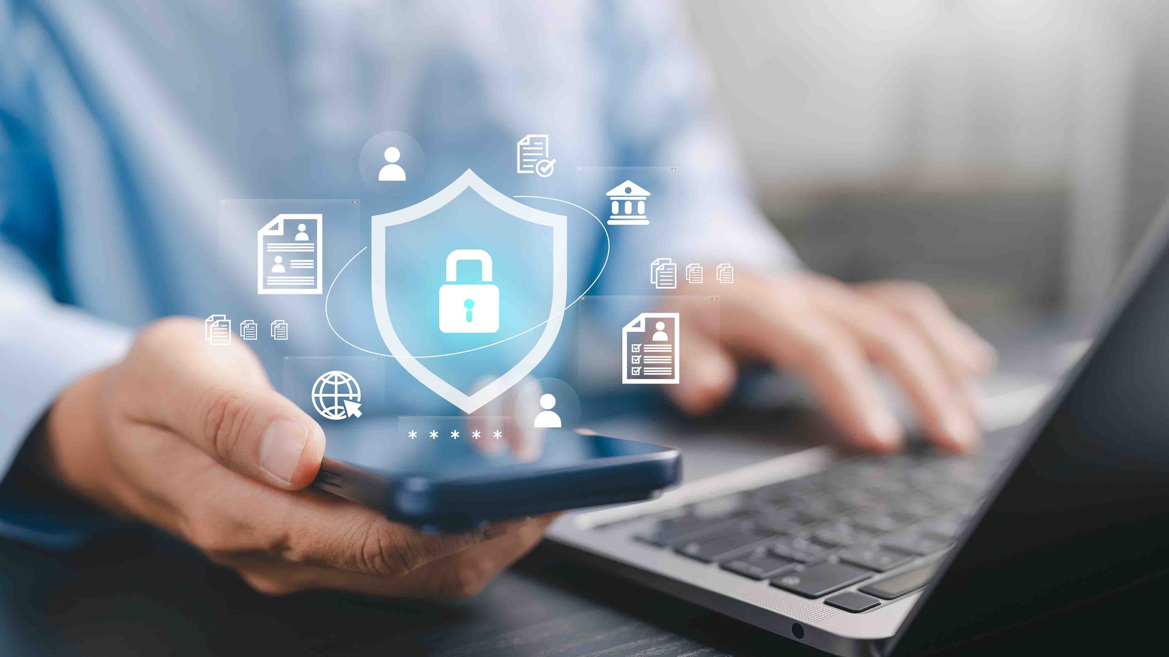 A person looks at their phone in their hand with their laptop open and security icons hover above the phone.