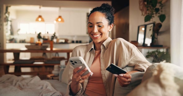 A woman sits on her couch in her living room using a credit card on her phone.