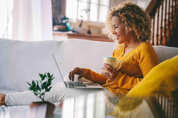 A female uses her computer while sipping a cup of coffee and sitting on a couch.