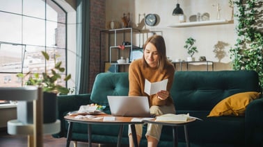 A woman sits on her green couch with her computer open looking at a piece of paper.