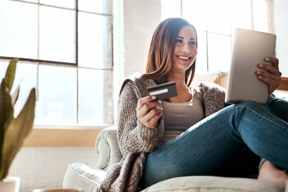 A woman sits on her couch with a debit card in her hand while on her tablet.
