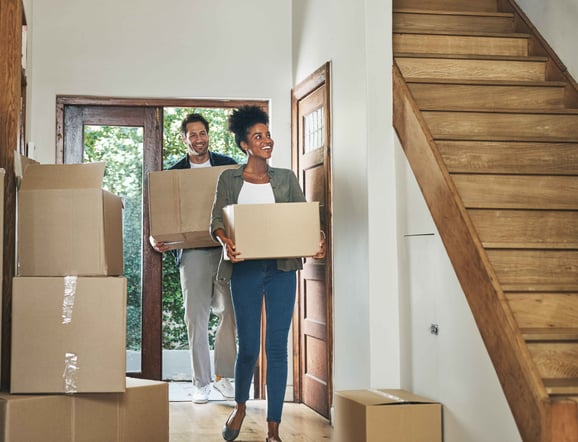 A young male and female walk into a home carrying boxes.