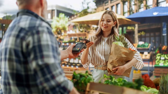 Female uses tap to pay at farmers market with groceries in her hand.