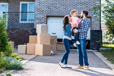 A man, woman, and child stand outside house with moving boxes in background.