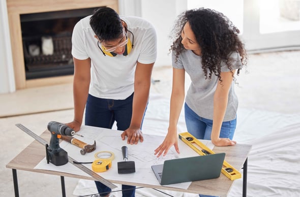 A man and woman stand at table with home improvement plans, computer, and tools.